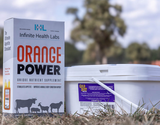 A Gold Power Pack box and a white Gold Dust tub from Infinite Health Labs sit on dry grass outdoors, with blurred trees and sky behind. The box shows animal silhouettes and highlights its benefits as a show animal supplement and appetite booster.